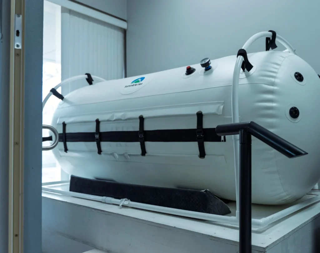 A white soft-sided lie-down hyperbaric chamber with a handrail beside it at K Wellness
