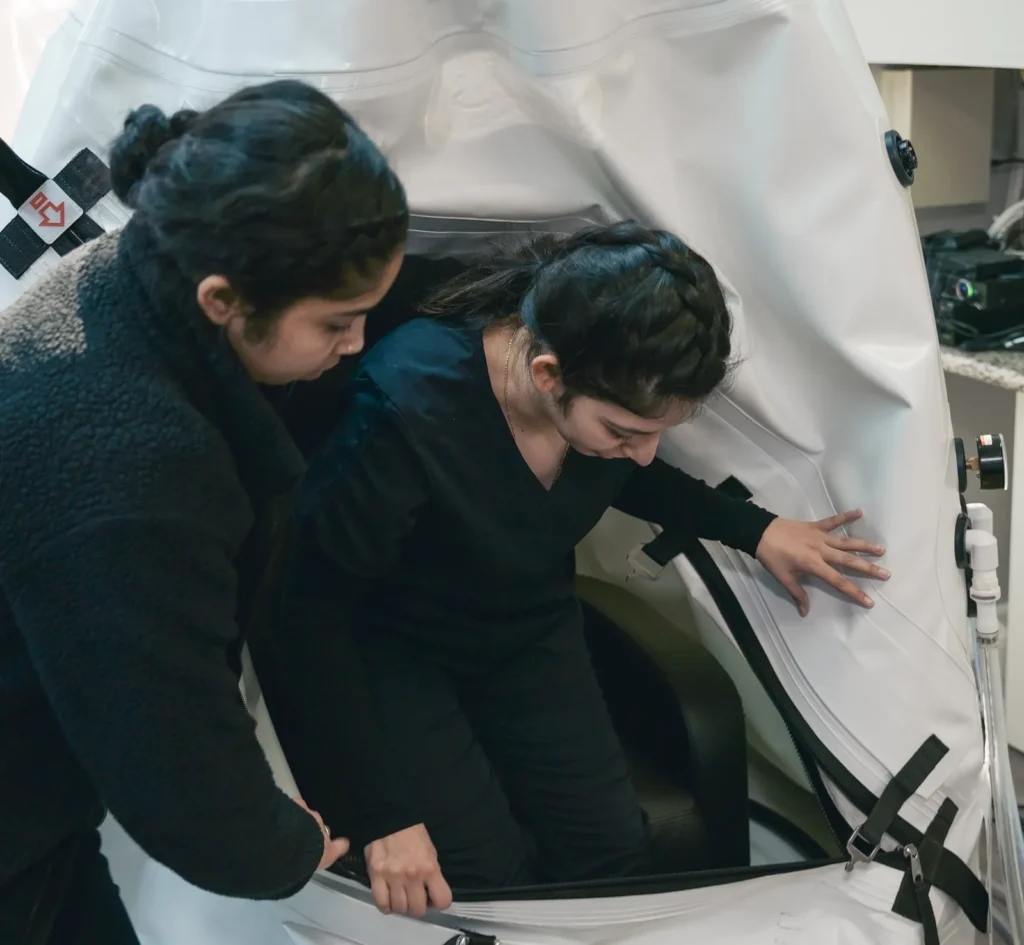 A K Wellness staff member helping someone into the sit-up hyperbaric chamber